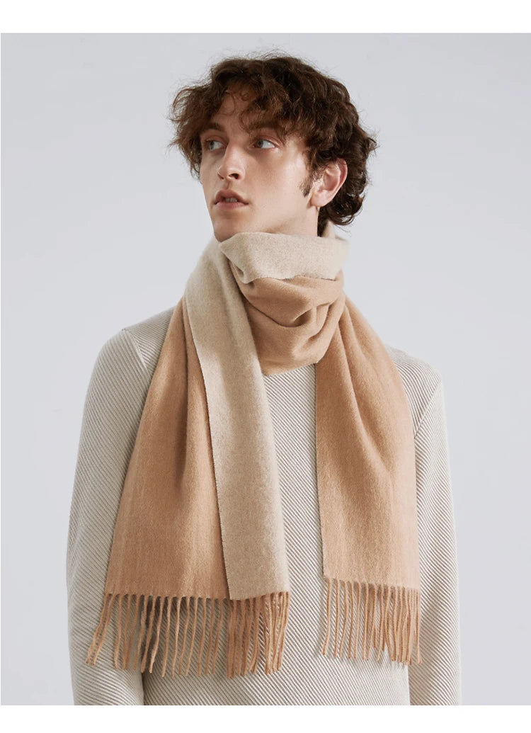 A man wearing a light beige knit sweater and a matching beige scarf with fringe, photographed against a plain light gray studio background.