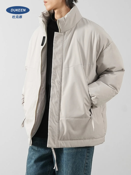 A person wearing a light beige puffer jacket with a stand-up collar, zippered front, and side pockets, layered over a black top and blue jeans. The brand logo 'DUKEEN' with Chinese characters is visible in the top left corner.