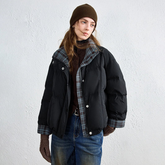 A model wearing a black puffer jacket with a gray plaid lining, a brown beanie, and blue jeans, standing against a textured white wall.
