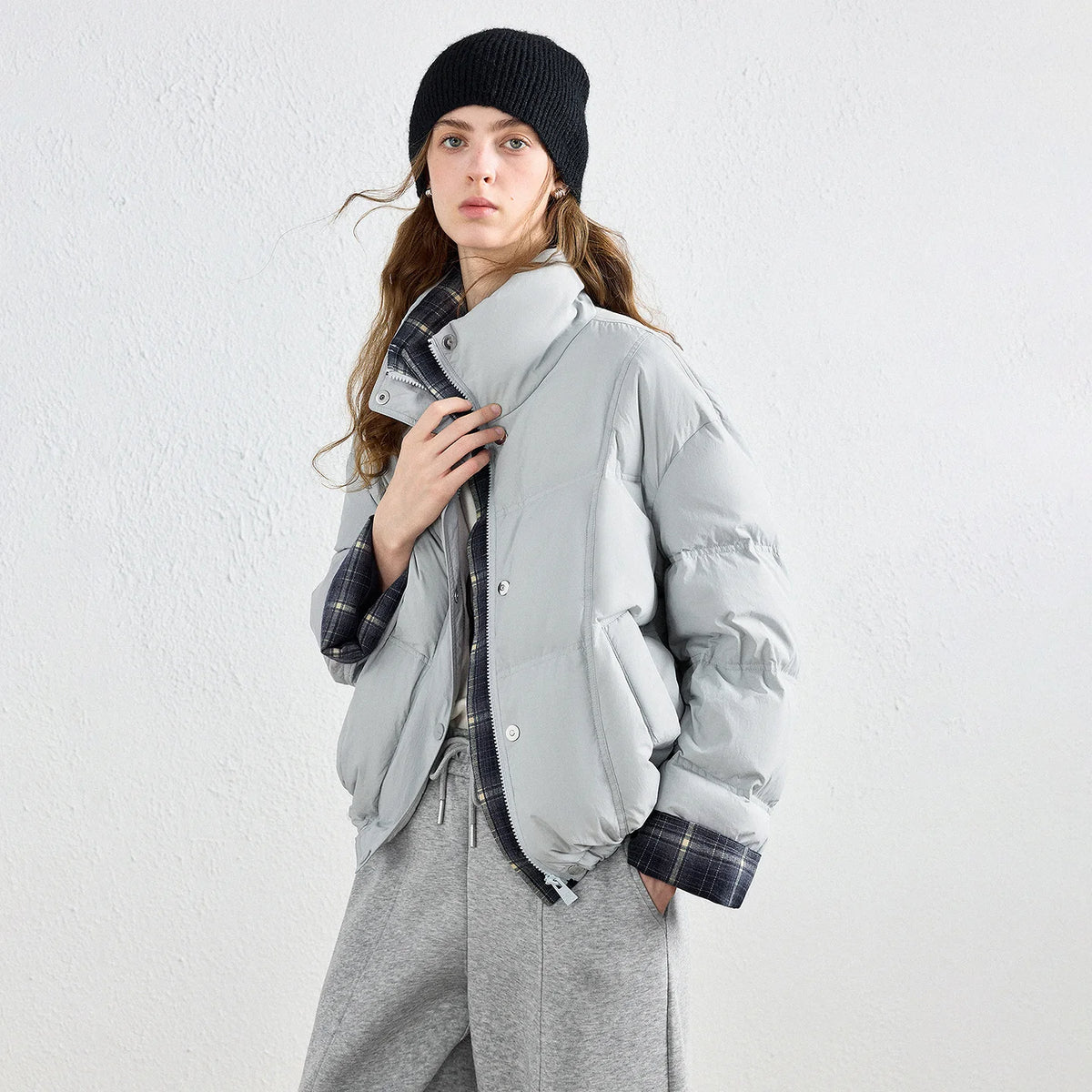 A woman wearing a light gray puffer jacket with a high collar and a visible plaid lining, styled with a black beanie, gray sweatpants, and a plaid shirt against a textured white wall.