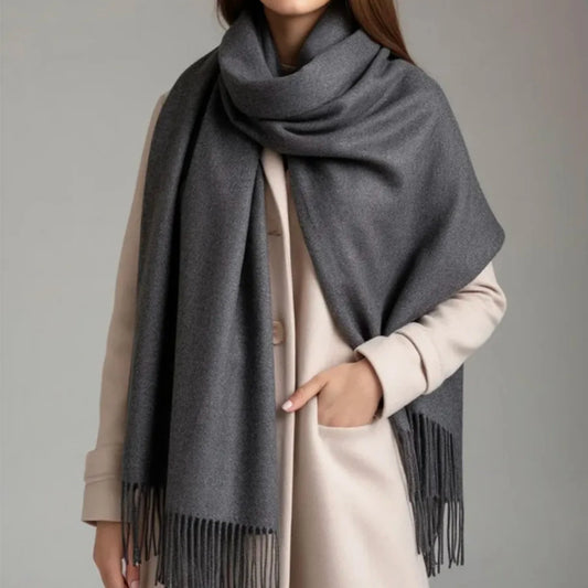 A woman wearing a large dark gray woolen shawl with fringe trim over a light beige coat, shown from the neck down against a plain light gray studio background.