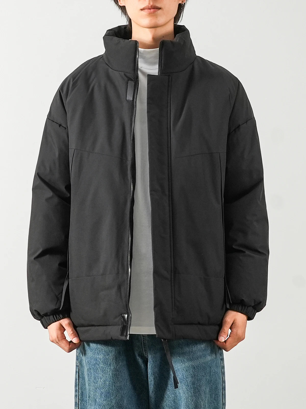 A person wearing a black puffer jacket with a high stand collar, front zipper, and zip pockets, layered over a gray shirt and blue jeans, photographed against a light gray studio background.