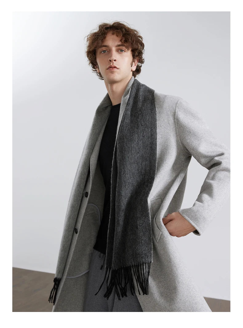 A man wearing a light gray wool-blend coat over a black shirt and a dark gray fringed scarf, standing against a plain light gray studio background.