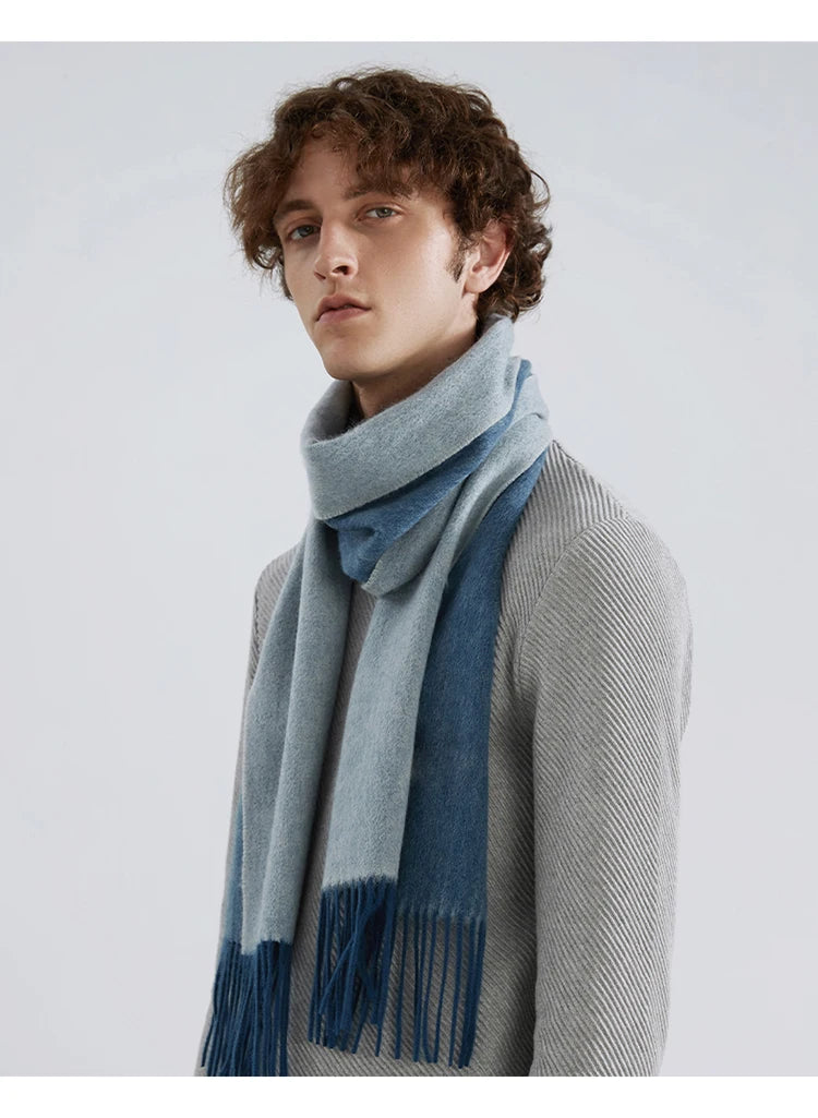 A man wearing a light blue and medium blue striped knit scarf with fringed ends, styled over a gray ribbed sweater against a light gray studio background.