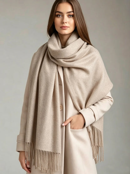 A woman wearing a large, soft beige scarf with fringe trim draped over a light beige coat against a neutral gray studio background.