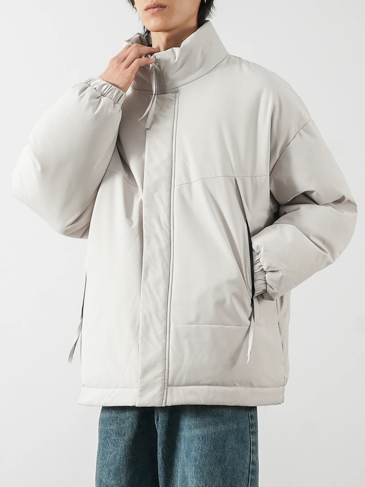 A person wearing an oversized light gray puffer jacket with a high collar and drawstring details, paired with blue jeans, against a plain light gray background.