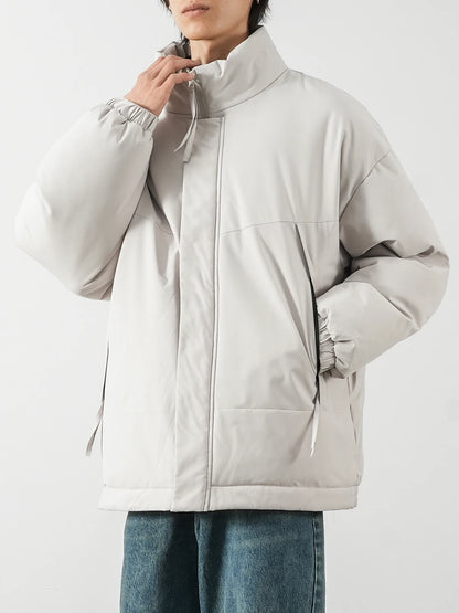 A person wearing an oversized light gray puffer jacket with a high collar and drawstring details, paired with blue jeans, against a plain light gray background.