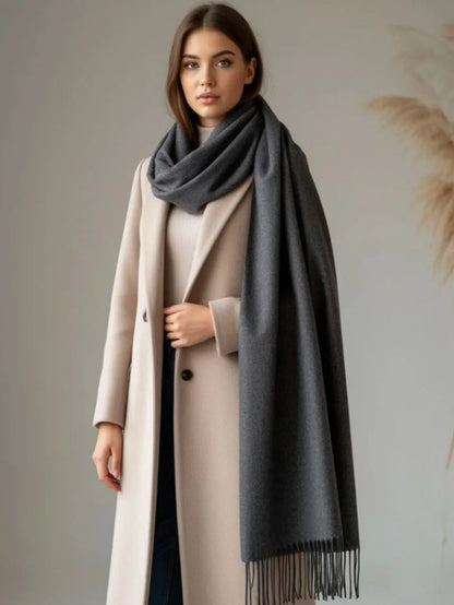 A woman wearing a long beige coat and a large dark gray scarf, standing against a soft neutral wall with dried grass decor in the background.