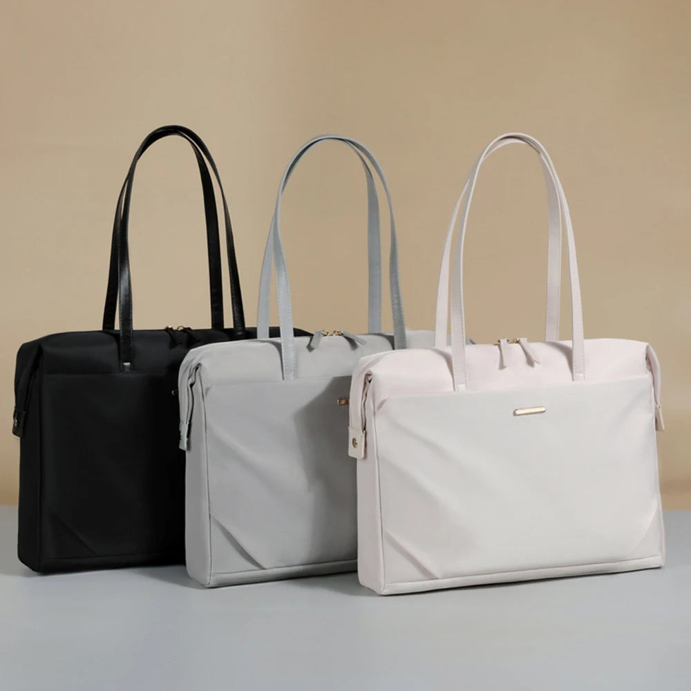 Three tote-style briefcase bags in black, light gray, and off-white are arranged side by side on a light gray surface against a warm beige background.