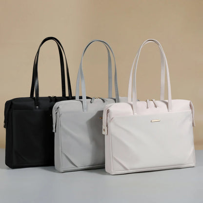 Three tote-style briefcase bags in black, light gray, and off-white are arranged side by side on a light gray surface against a warm beige background.