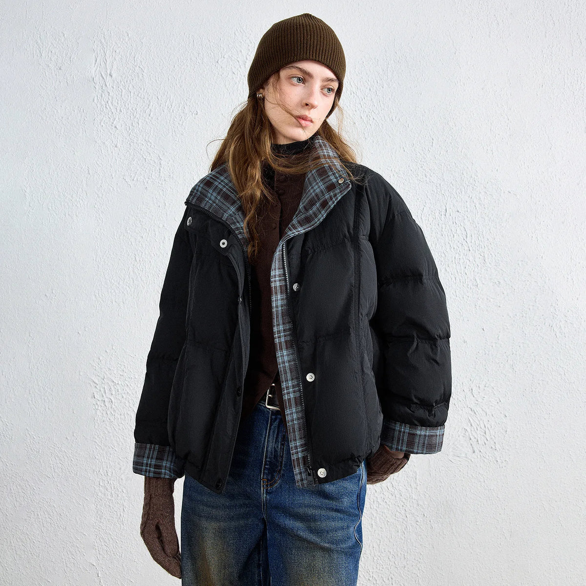 A model wearing a black puffer jacket with a gray plaid lining, a brown beanie, and blue jeans, standing against a textured white wall.