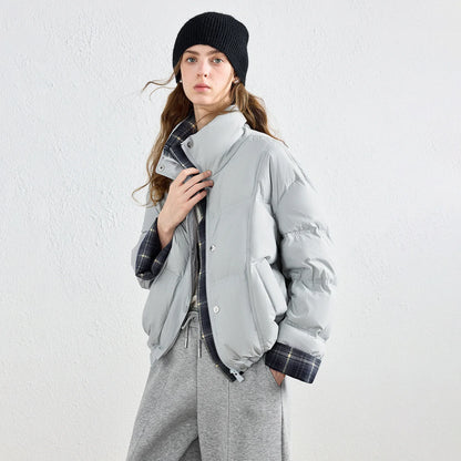 A woman wearing a light gray puffer jacket with a high collar and a visible plaid lining, styled with a black beanie, gray sweatpants, and a plaid shirt against a textured white wall.