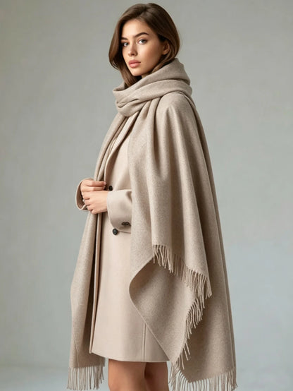 A woman wearing a light beige double-breasted coat with a matching fringed wrap shawl draped over her shoulders, posed against a neutral gray studio background.