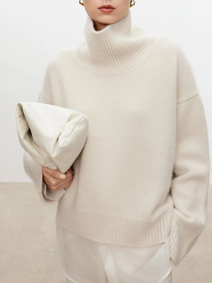 Person wearing a beige turtleneck sweater holding a matching clutch against a white background