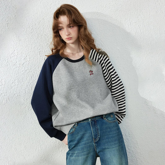 A woman wearing a raglan-style long-sleeve sweatshirt with a heather gray body, navy blue sleeves, and black-and-white striped sleeves, paired with blue jeans, standing against a textured white wall.