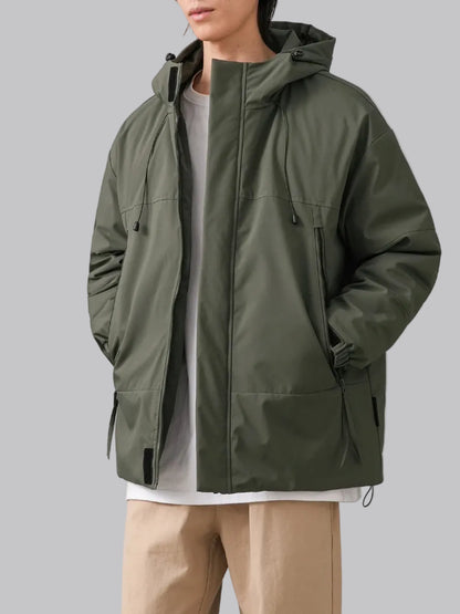 A man wearing an olive green hooded jacket by DUKEEEN over a white t-shirt and beige pants, shown against a light gray studio background with a product info badge on the left.