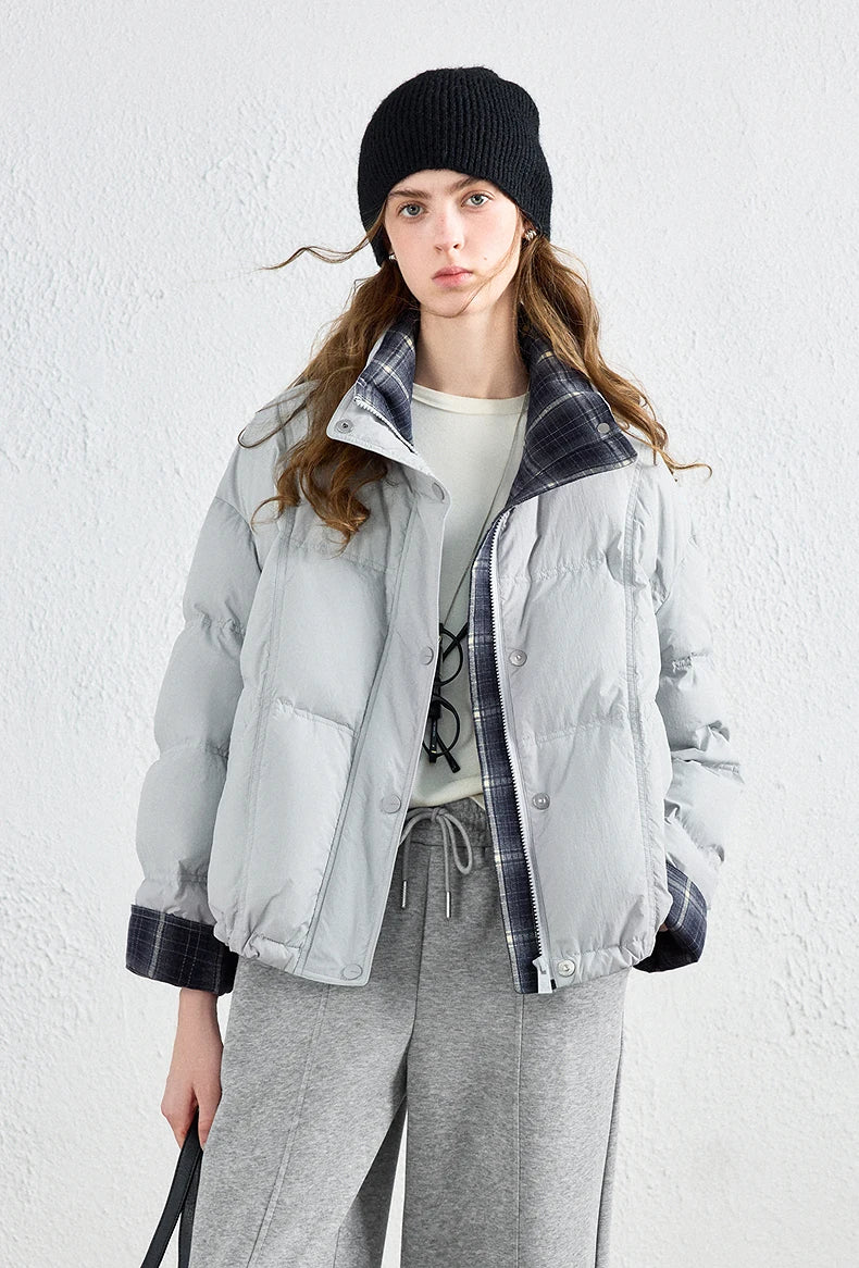 A model wearing a light gray puffer jacket with a dark gray and black plaid collar and cuffs, styled with gray sweatpants, a white t-shirt, and a black beanie against a textured white wall.