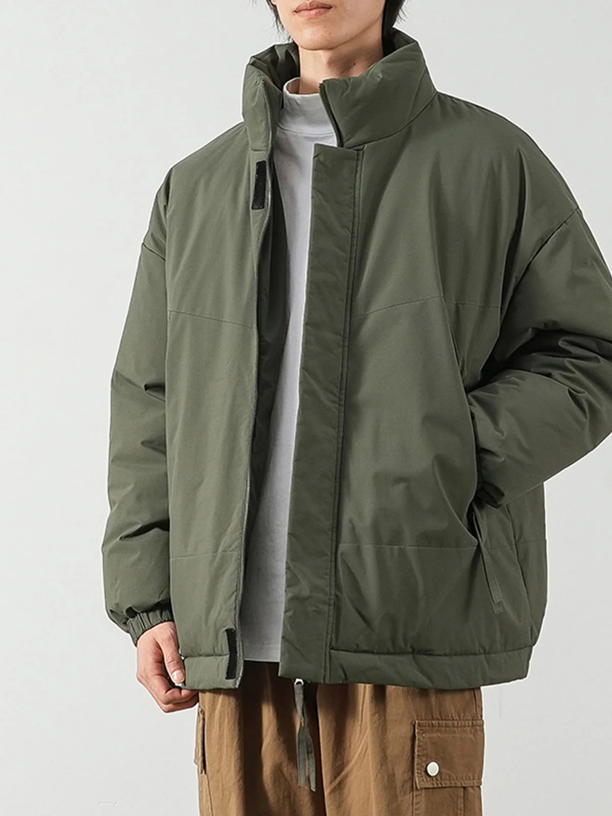 A person wearing an oversized olive green jacket over a light gray shirt and brown pants, photographed against a plain light gray studio background.