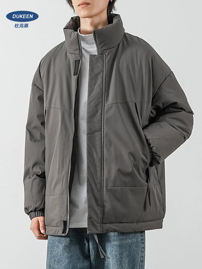 A person wearing an oversized dark gray puffer jacket over a light gray shirt and blue jeans, shown from the neck down against a plain light gray studio background. A DUKEEN logo appears in the top-left corner.