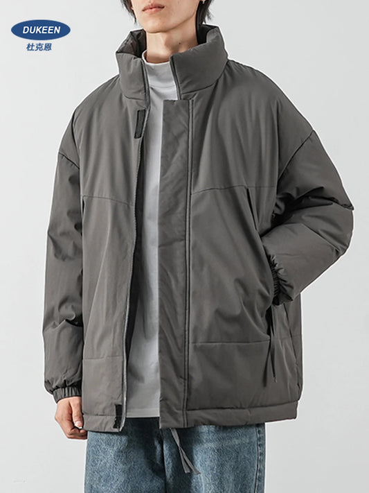 A person wearing an oversized dark gray puffer jacket over a light gray shirt and blue jeans, shown from the neck down against a plain light gray studio background. A DUKEEN logo appears in the top-left corner.