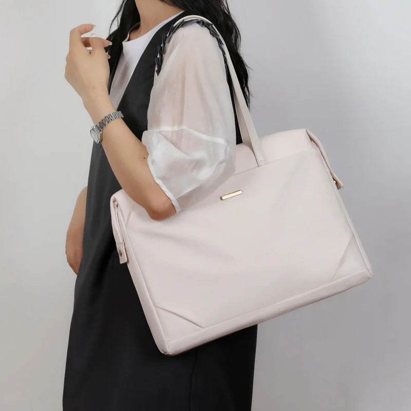 A woman wearing a black and white dress carries a large, structured light pink tote bag with a gold-tone logo plate on a plain light gray background.