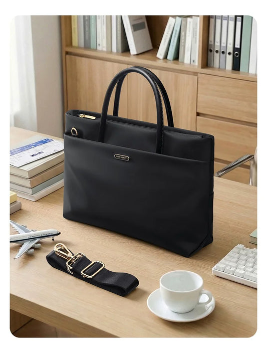 A black structured tote bag with dual top handles sits on a light wood desk, with an additional detachable black shoulder strap placed in front. The scene includes a white coffee cup, books, a keyboard, and a wooden bookshelf in the background, indicating a lifestyle office setting.