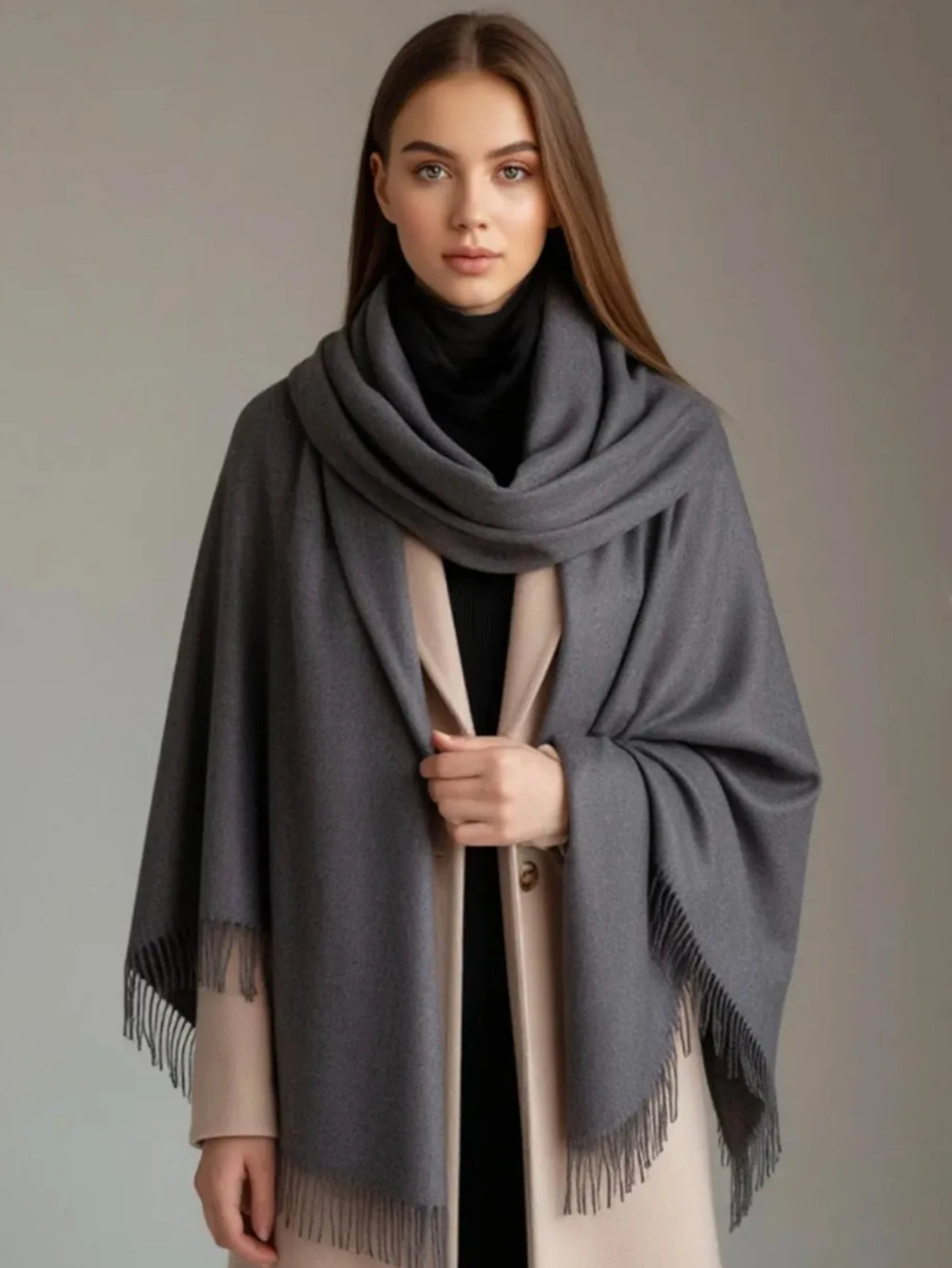 A woman modeling a large charcoal gray fringed wrap or shawl over a black turtleneck and beige coat against a plain gray studio background.
