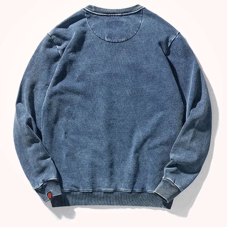 A washed blue long-sleeve crewneck sweatshirt shown flat against a light off-white background. The fabric has a faded, mineral-washed texture, and there is a small red tag on the left cuff.