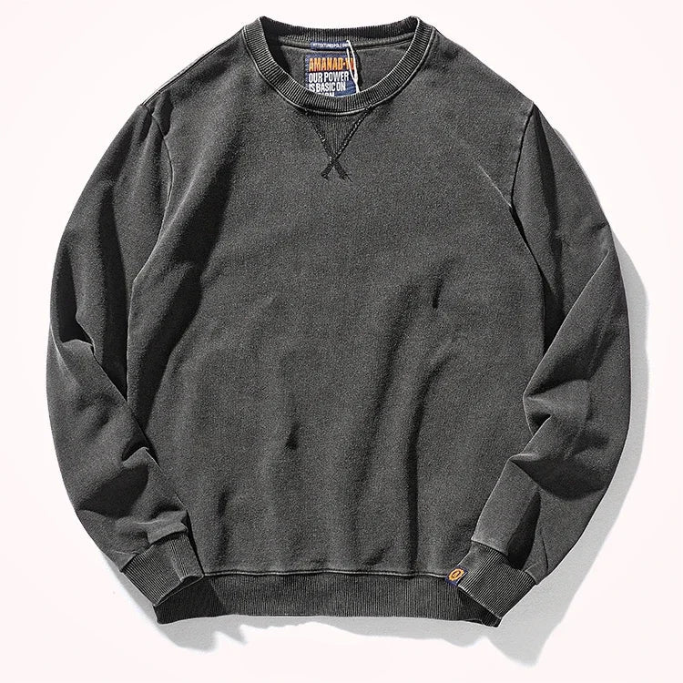 A dark charcoal gray crewneck sweatshirt laid flat on a light off-white background. The sweatshirt features a classic crew neck, ribbed cuffs, and a ribbed hem, with a small orange tag near the bottom right cuff.