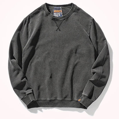 A dark charcoal gray crewneck sweatshirt laid flat on a light off-white background. The sweatshirt features a classic crew neck, ribbed cuffs, and a ribbed hem, with a small orange tag near the bottom right cuff.