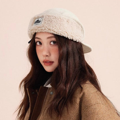 Woman wearing a beige sherpa-lined cap and brown coat against a light pink background